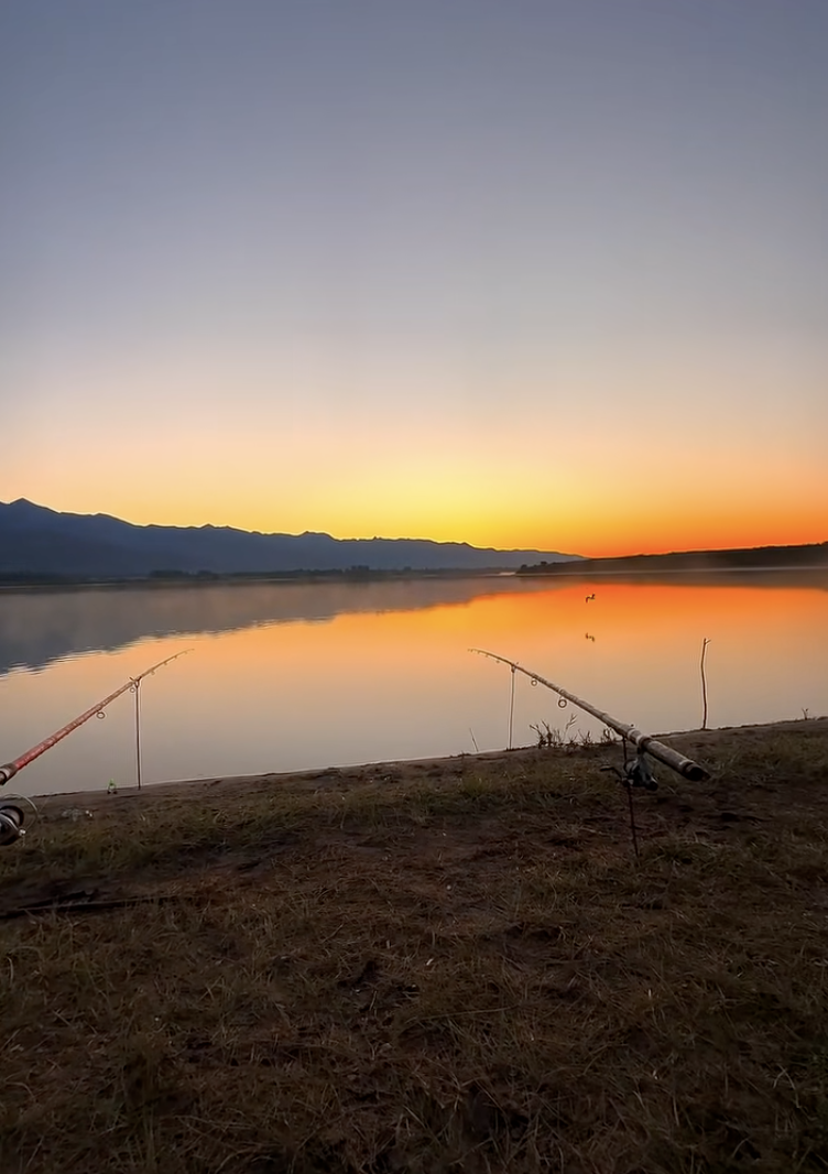 Duas varas de pesca na margem de um lago, com montanhas ao fundo e o reflexo de um céu alaranjado na água.