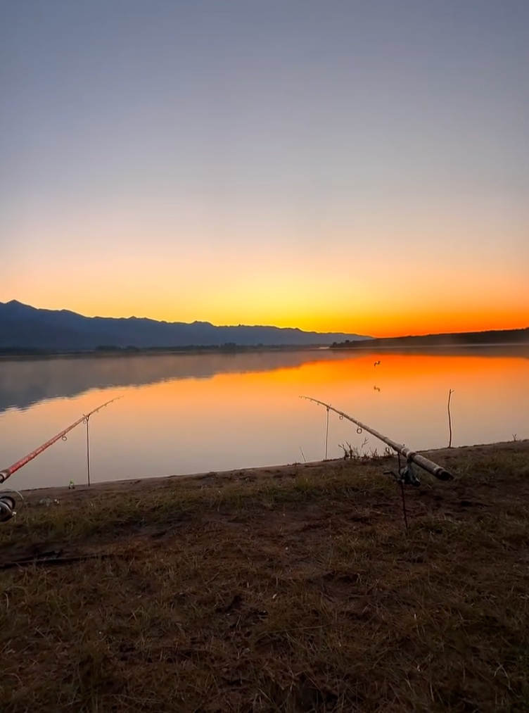 Duas varas de pesca à beira de águas calmas que refletem o céu alaranjado, com montanhas no horizonte.